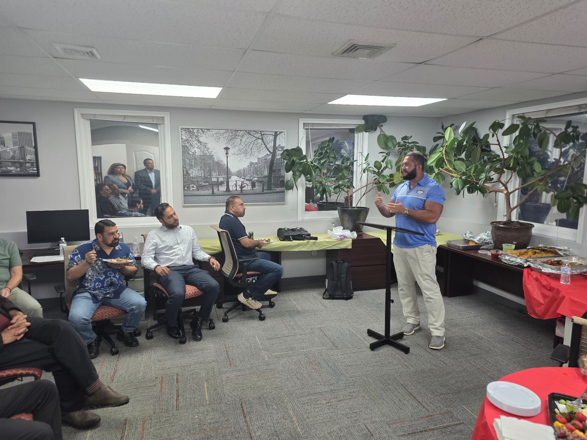 Speaker at a podium addressing attendees seated in an office meeting room during a lunch-and-learn session